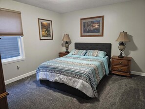 Quarto com cama, duas mesas de cabeceira, duas luminárias, janela com persianas e duas imagens emolduradas na parede.