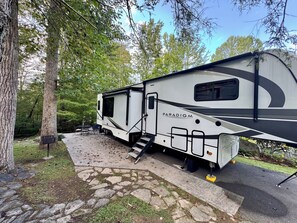 Un camping-car blanc, arborant le logo « PARADIGM », est stationné sur un terrain de camping. Un sentier pavé de pierres mène à une table et à des chaises.