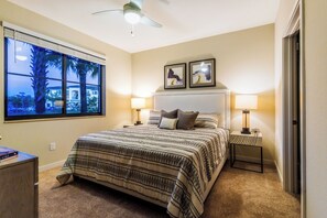 Un dormitorio con una cama grande, dos lámparas de noche, un ventilador de techo y una ventana con vista a palmeras y edificios.