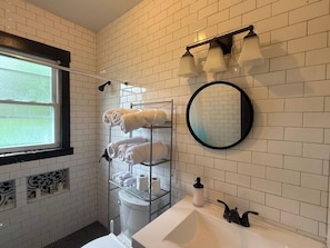 Ein Badezimmer mit weißen Fliesen, einem Fenster, einem Handtuchhalter, einer Toilette, einem Waschbecken, einem Spiegel und einer Dusche.