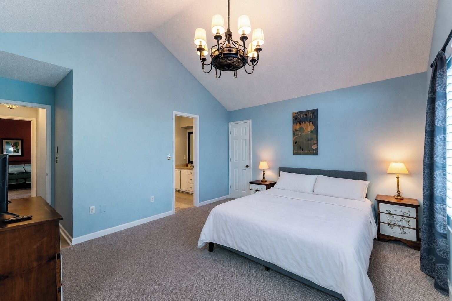 A bedroom with a bed, a chandelier, a nightstand, a lamp, and a painting on the wall.