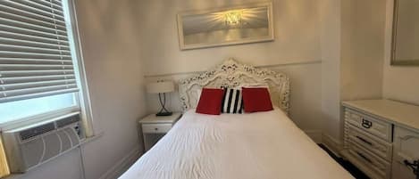 A hotel room with a bed, a bedside table with a lamp, a dresser, and a window with blinds.