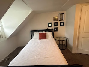Ein Schlafzimmer mit einem Bett, einem kleinen runden Tisch, einer Tür und einer schrägen Decke mit Oberlicht.