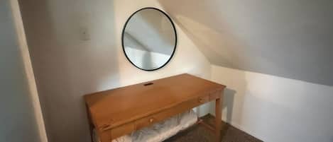 A small, single-bed room with a wooden bench, a round mirror on the wall, and a carpeted floor.