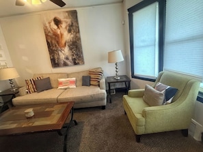 Ein Wohnzimmer mit einem beigen Sofa, einem grünen Sessel, einem Couchtisch, einer Lampe, einem Bild an der Wand und einem Fenster mit Jalousien.