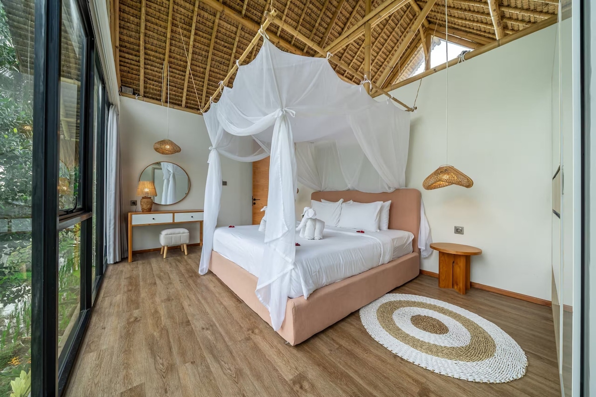 A bedroom with a canopy bed, bedside tables, a mirror, and a view of greenery outside.