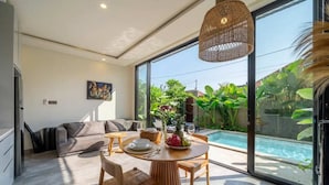 A modern living space with a dining area, a sofa, and a large window overlooking a swimming pool and lush greenery. - Green Harmony Villas (Gianyar)