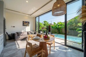 A modern living space with a dining area, a sofa, and a large window overlooking a swimming pool and lush greenery. - Green Harmony Villas (Gianyar)