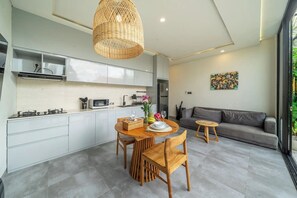Una cocina moderna con armarios blancos, un comedor con sillas de madera, un sofá gris y una lámpara colgante de mimbre.