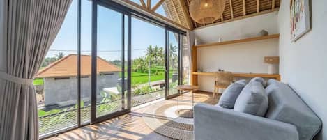 A modern living room with a large sliding glass door leading to a garden, a sofa, a small table, and a hanging light fixture.