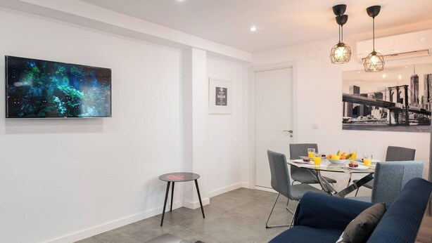 Ein modernes Wohnzimmer mit einem blauen Sofa, einem Esstisch mit Stühlen, einem kleinen runden Tisch und einem großen Flachbildfernseher an der Wand.
