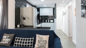 Ein modernes Wohnzimmer mit einem dunkelblauen Sofa, zwei Zierkissen, einem Schreibtisch mit Lampe und Blick in einen Küchenbereich mit weißen Schränken und einer Mikrowelle.
