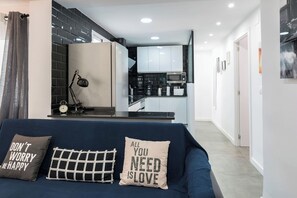 Uma sala de estar moderna com um sofá azul escuro, duas almofadas decorativas, uma escrivaninha com abajur e vista para uma área de cozinha com armários brancos e micro-ondas.