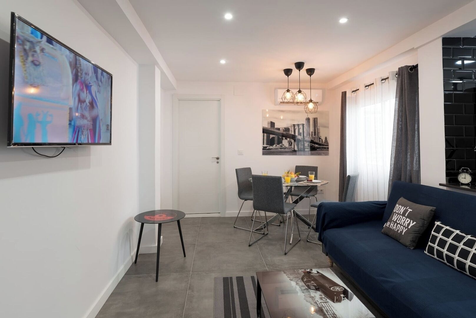 A modern living room with a blue sofa, a dining area with chairs, a small table, and a TV mounted on the wall.
