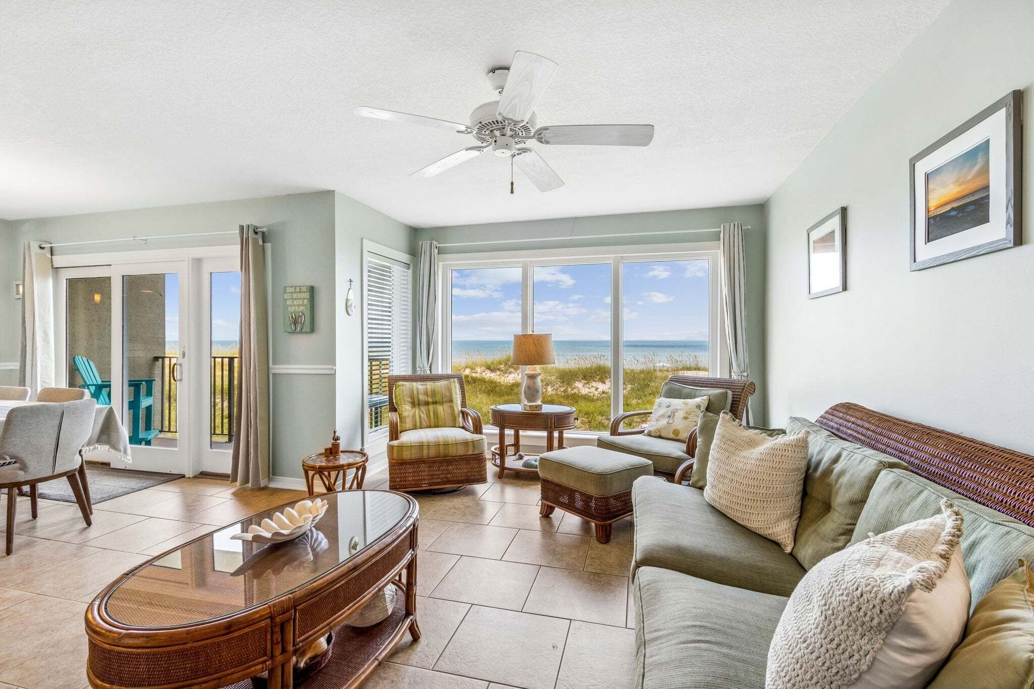 A spacious living room with a view of the beach, a ceiling fan, and a dining area with a glass-top table.