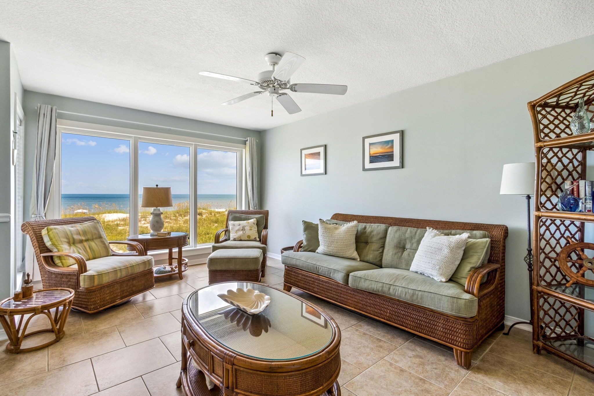 Un salon spacieux doté d’une grande fenêtre donnant sur la plage, d’un ventilateur de plafond, de meubles en osier et d’une table basse en verre.