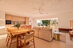 Eine moderne Küche mit Holzschränken, ein Essbereich mit Holztisch und Stühlen, eine Sitzecke und ein großes Fenster mit Blick ins Freie.