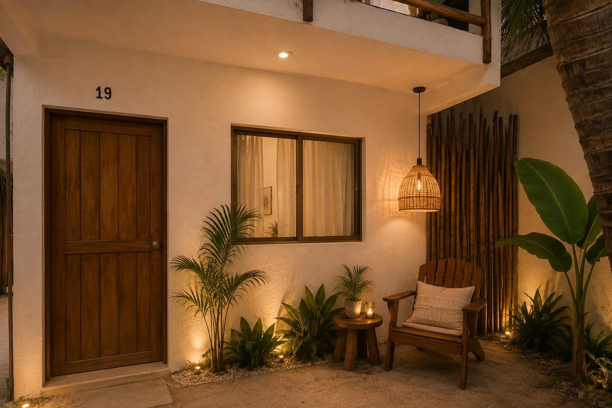 A building entrance with a wooden door, a window with curtains, a wicker lamp, a chair, and various plants.
