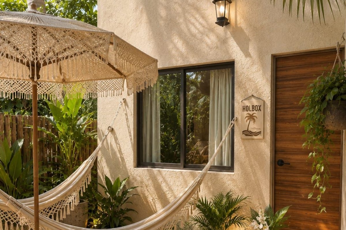 A hammock, a wooden table, wicker chairs, and a sign reading 'HOLBOX' in a courtyard with plants and a building in the background.