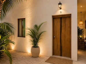A building entrance with a wooden double door, a potted plant, a wall-mounted light fixture, and a number '5' sign. - Hotel Holbox Centro (Isla Holbox)