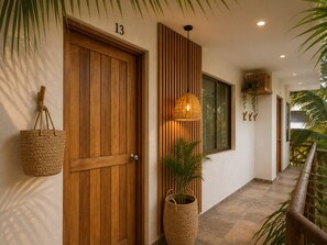 A hallway with a wooden door labeled '13', a woven basket hanging on the wall, a woven floor lamp, and a potted plant. - Hotel Holbox Centro (Isla Holbox)