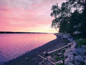 Une vue sur un lac, avec une rive rocheuse, des arbres et une étendue d’eau calme qui reflète le ciel.