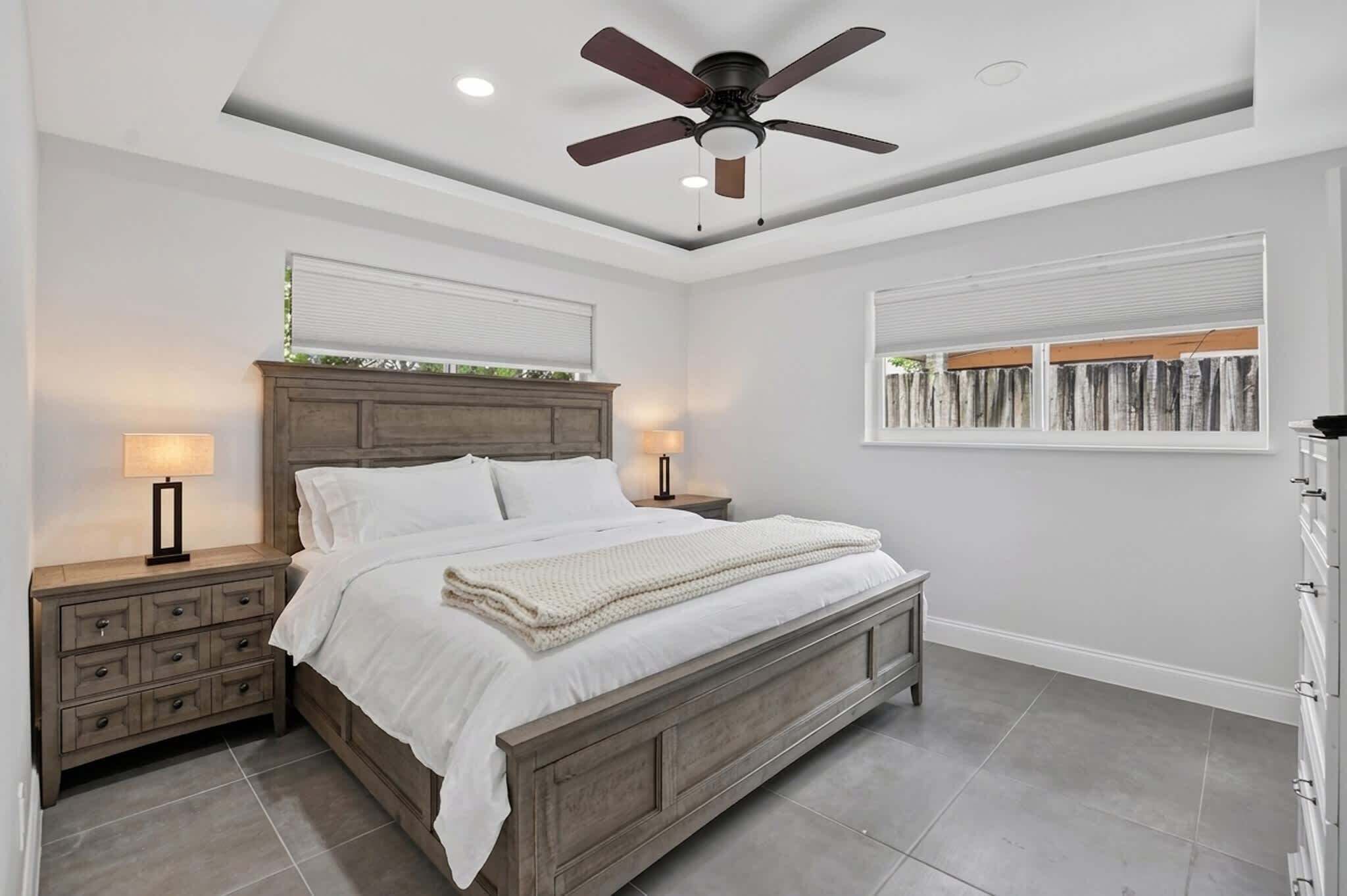 Une chambre à coucher comprenant un lit, des tables de chevet, un ventilateur de plafond et une fenêtre avec des stores.
