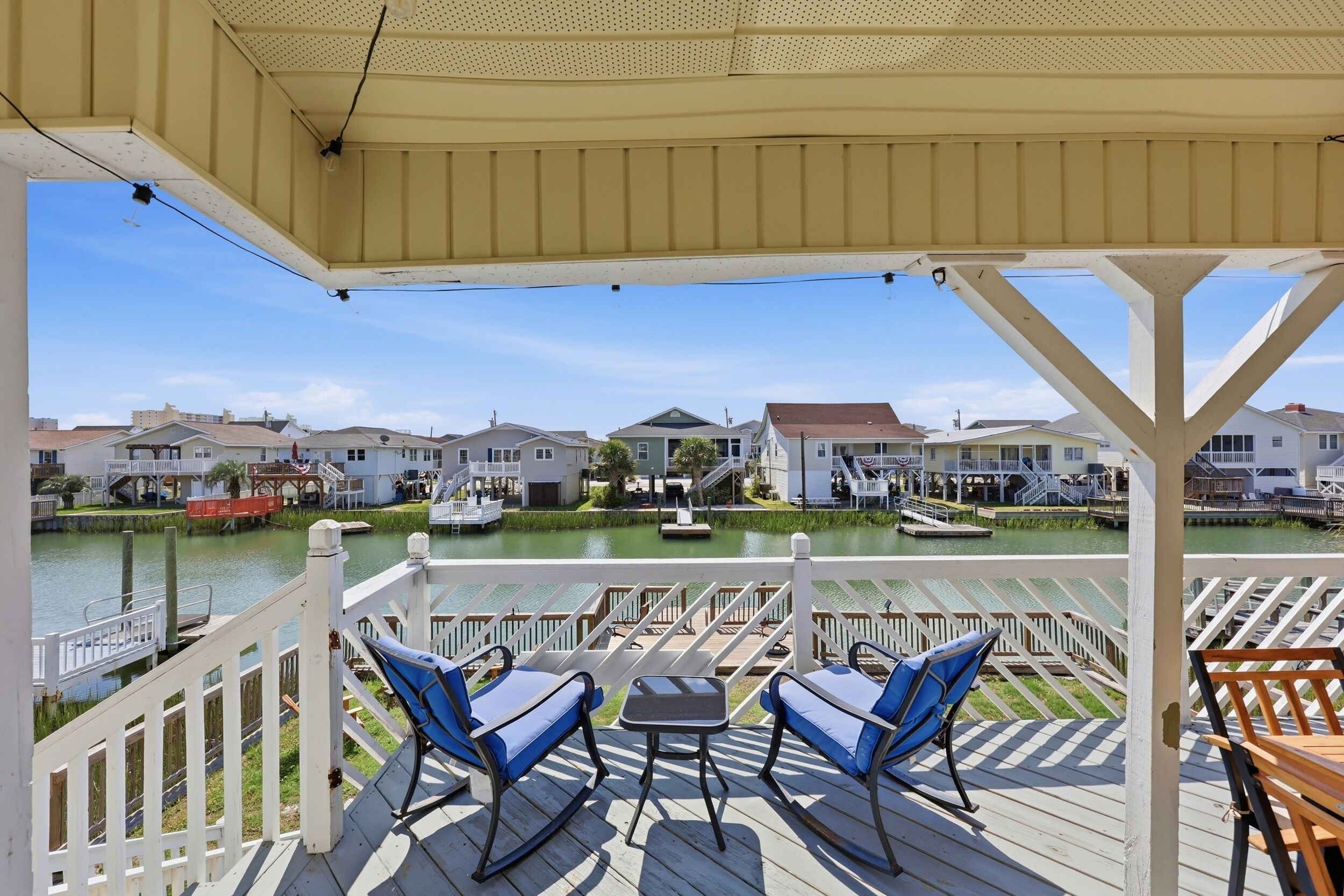 Une terrasse extérieure couverte, dotée de deux fauteuils à bascule et d’une petite table, qui offre une vue sur une étendue d’eau paisible, avec des maisons en arrière-plan.