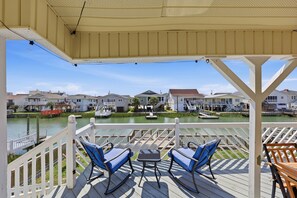 A covered outdoor deck with two rocking chairs and a small table, overlooking a calm water body with houses in the background. - Silver Sand Dollar | Upscale 6BR Escape-Hot Tub (North Myrtle Beach)