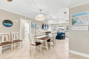 A spacious dining area with a large wooden table and white chairs, a kitchen with blue cabinets, and a beach-themed wall art. - Silver Sand Dollar | Upscale 6BR Escape-Hot Tub (North Myrtle Beach)