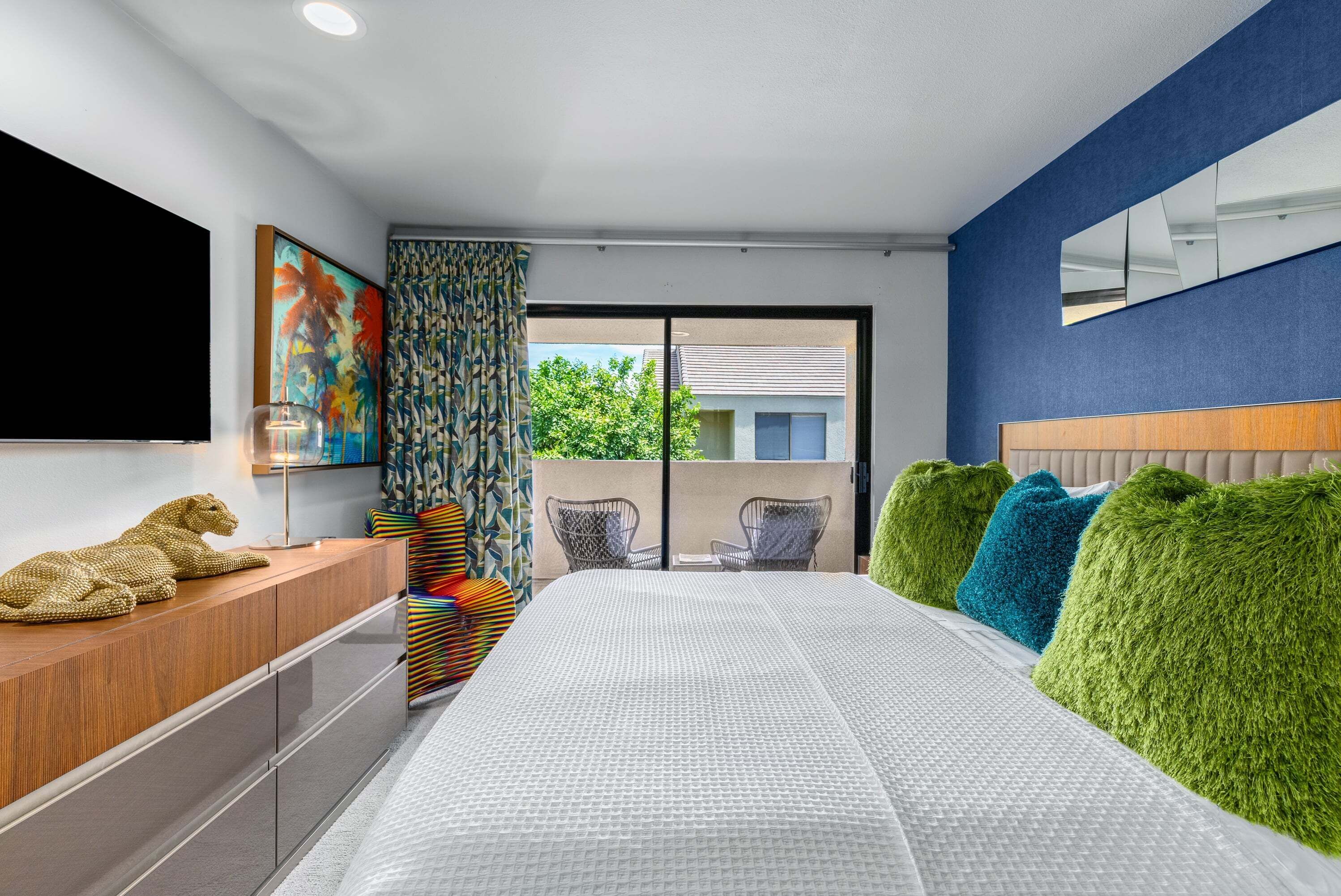 A modern bedroom with a large bed, a flat-screen TV, a wooden dresser, and a view of the outdoors.