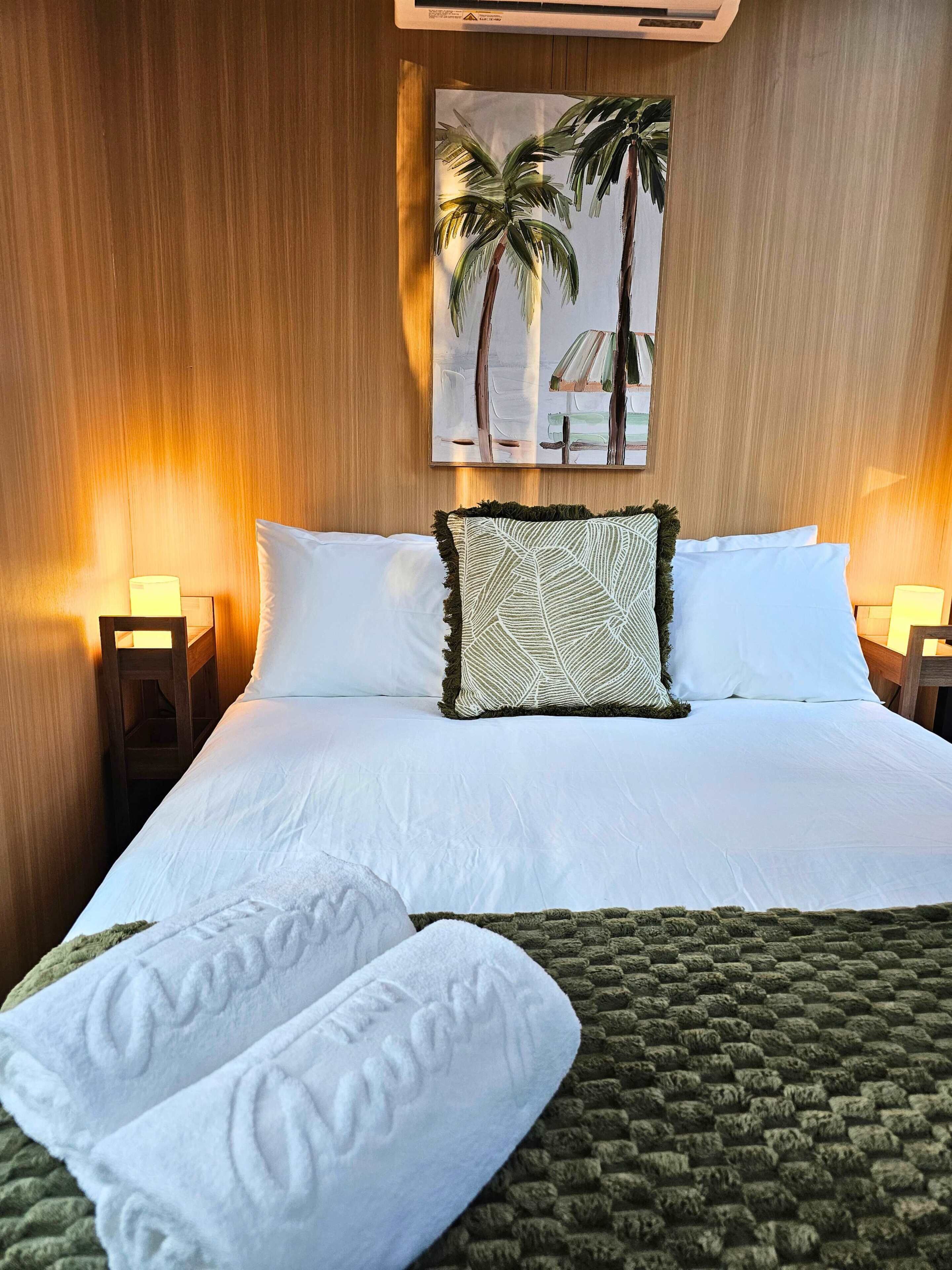 Een hotelkamer met een groot bed, twee bedlampen, een schilderij van palmbomen en een groen, gertextureerd plaid.