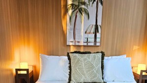 Habitació d'hotel amb un llit gran, dues làmpades de nit, un quadre de palmeres i una manta verda amb textura.