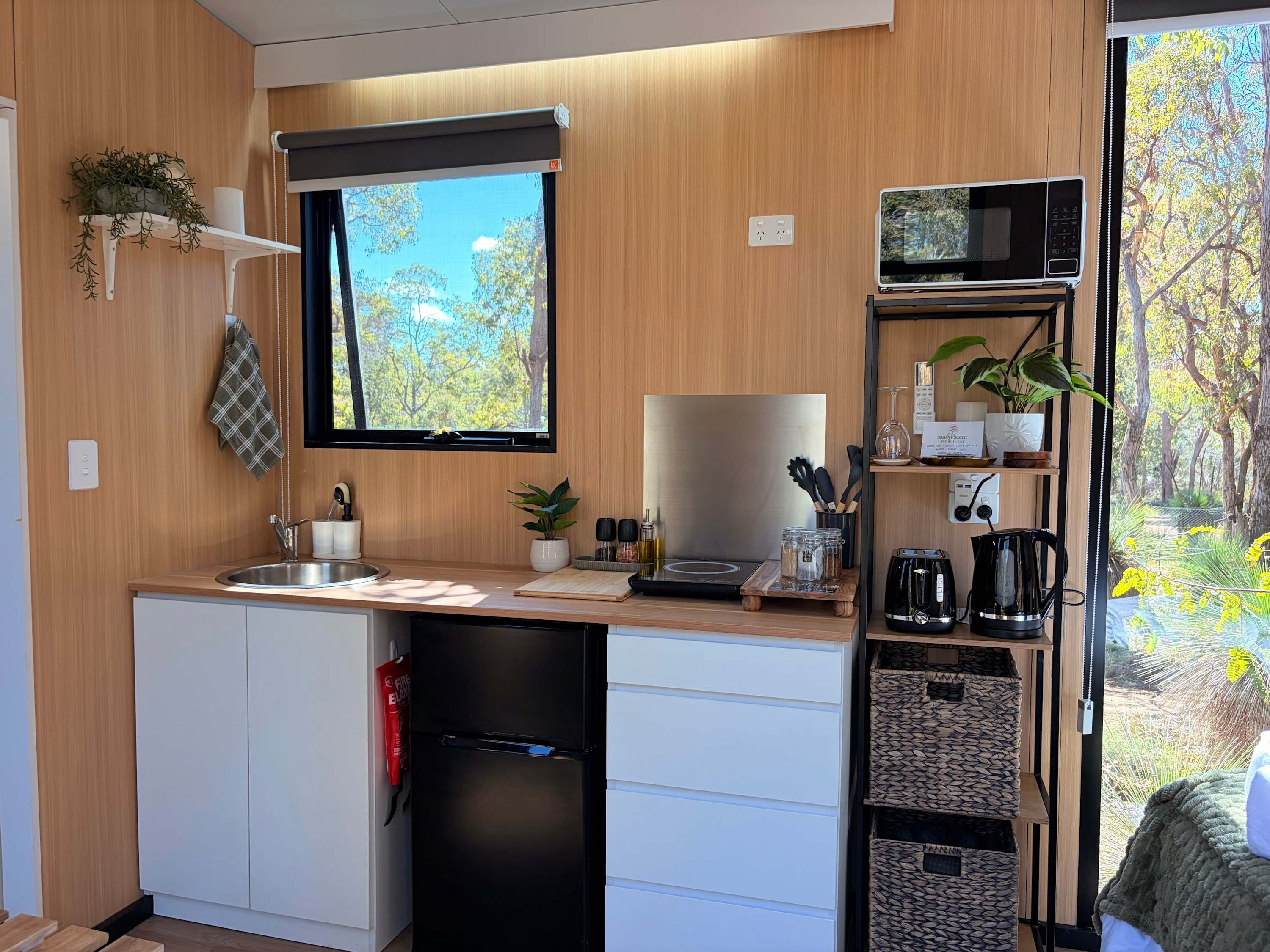 Cozinha compacta com micro-ondas integrado, pia e geladeira. Há uma janela com vista para árvores e uma prateleira com utensílios de cozinha.
