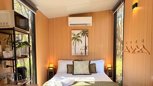 A hotel room with a bed, a nightstand, a shelf with kitchen items, a wall-mounted air conditioner, and a window with a view of palm trees.