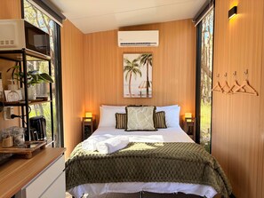 A hotel room with a bed, a nightstand, a shelf with kitchen items, a wall-mounted air conditioner, and a window with a view of palm trees.