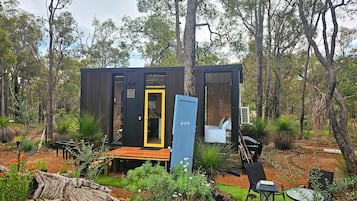 Een kleine, moderne blokhut met een terras, omgeven door een tuin en bomen.