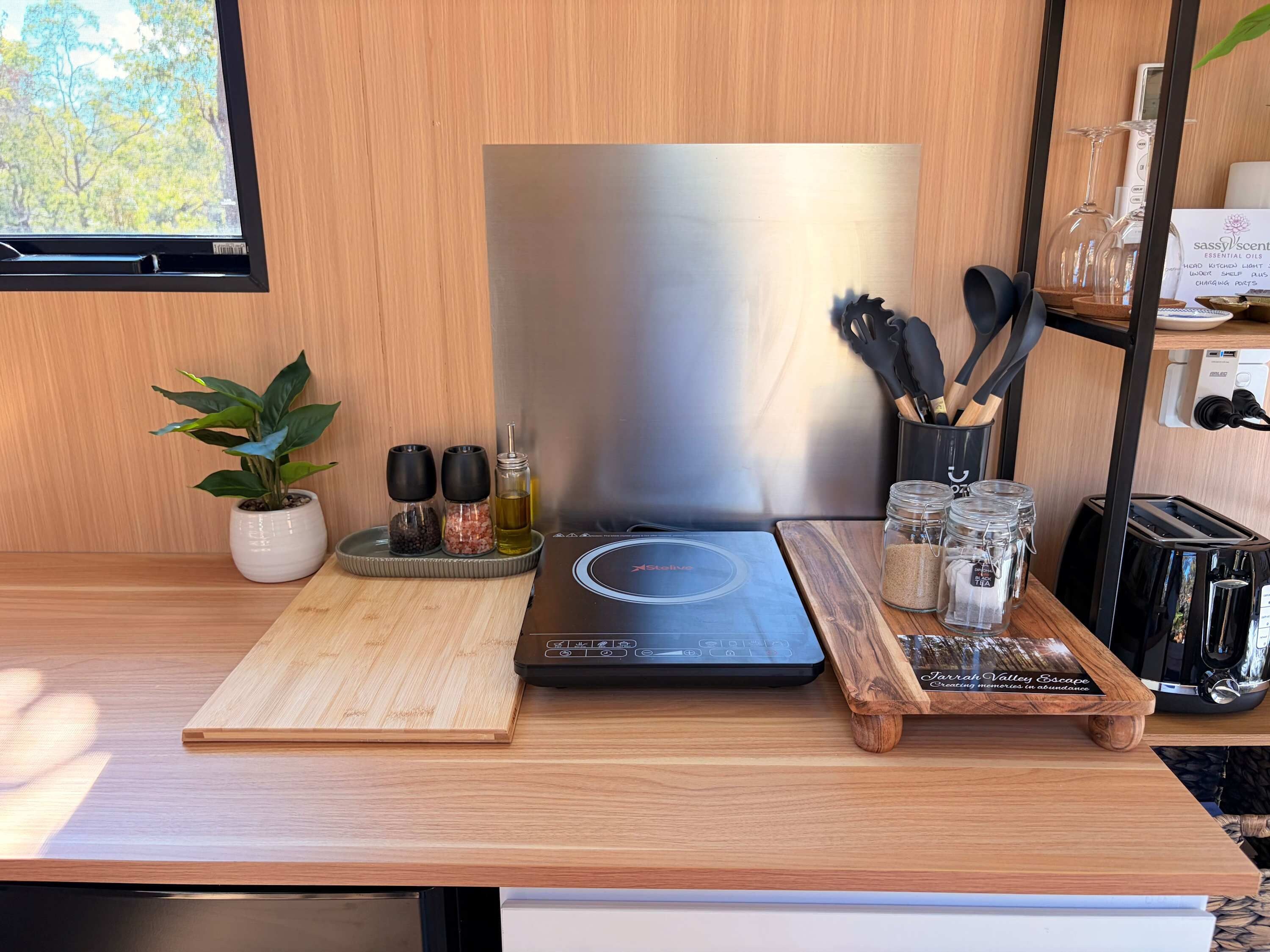 A kitchen counter with a stainless steel appliance, a potted plant, salt and pepper shakers, a cutting board, and a wooden tray with jars.