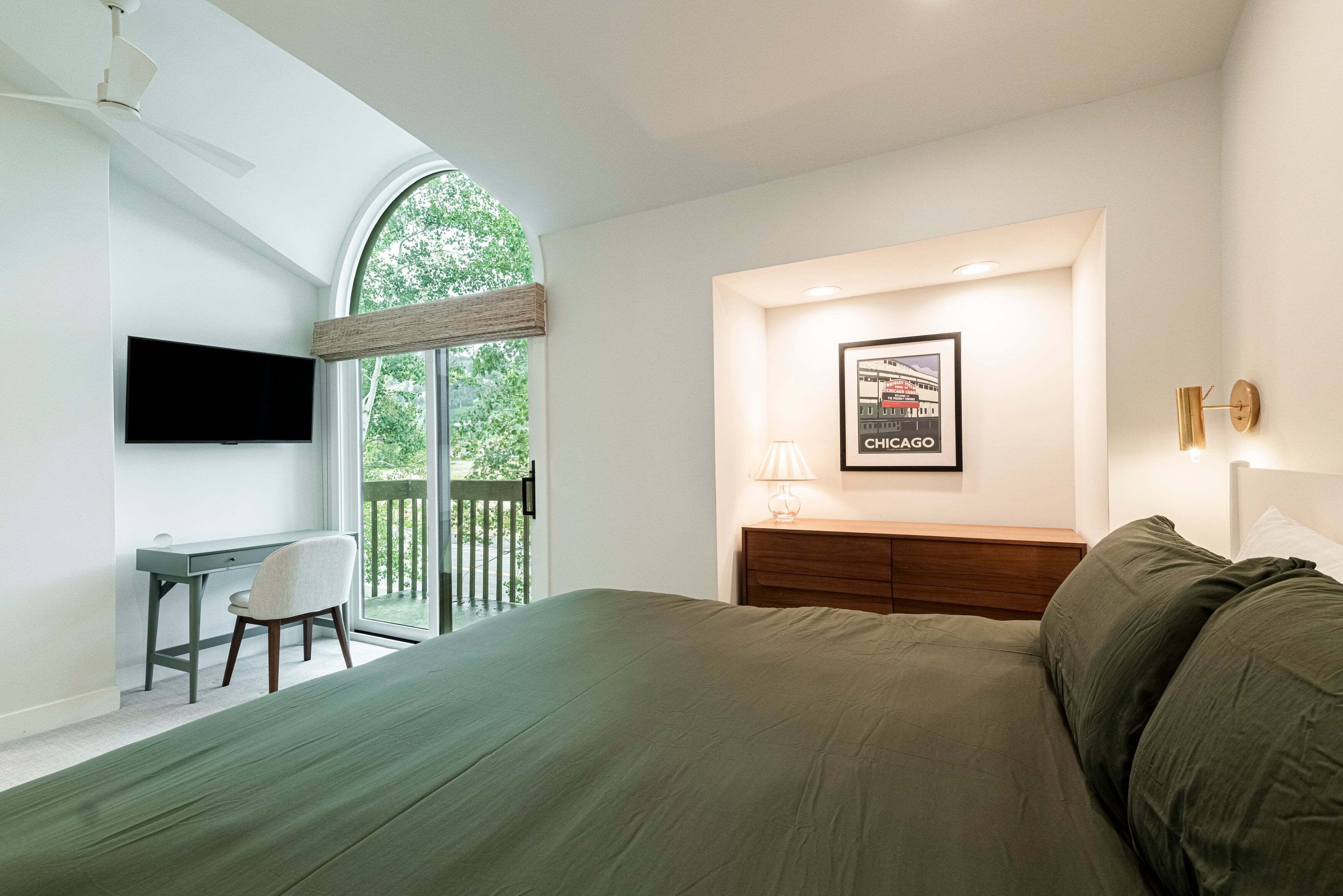 A bedroom with a large bed, a desk, and a chair. There is a window with a view of greenery outside.