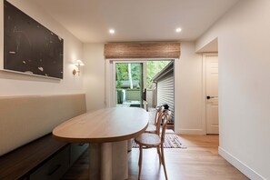 Interior moderno com uma mesa redonda, um banco e uma cadeira. Uma porta dá acesso a uma área externa com vista para árvores.