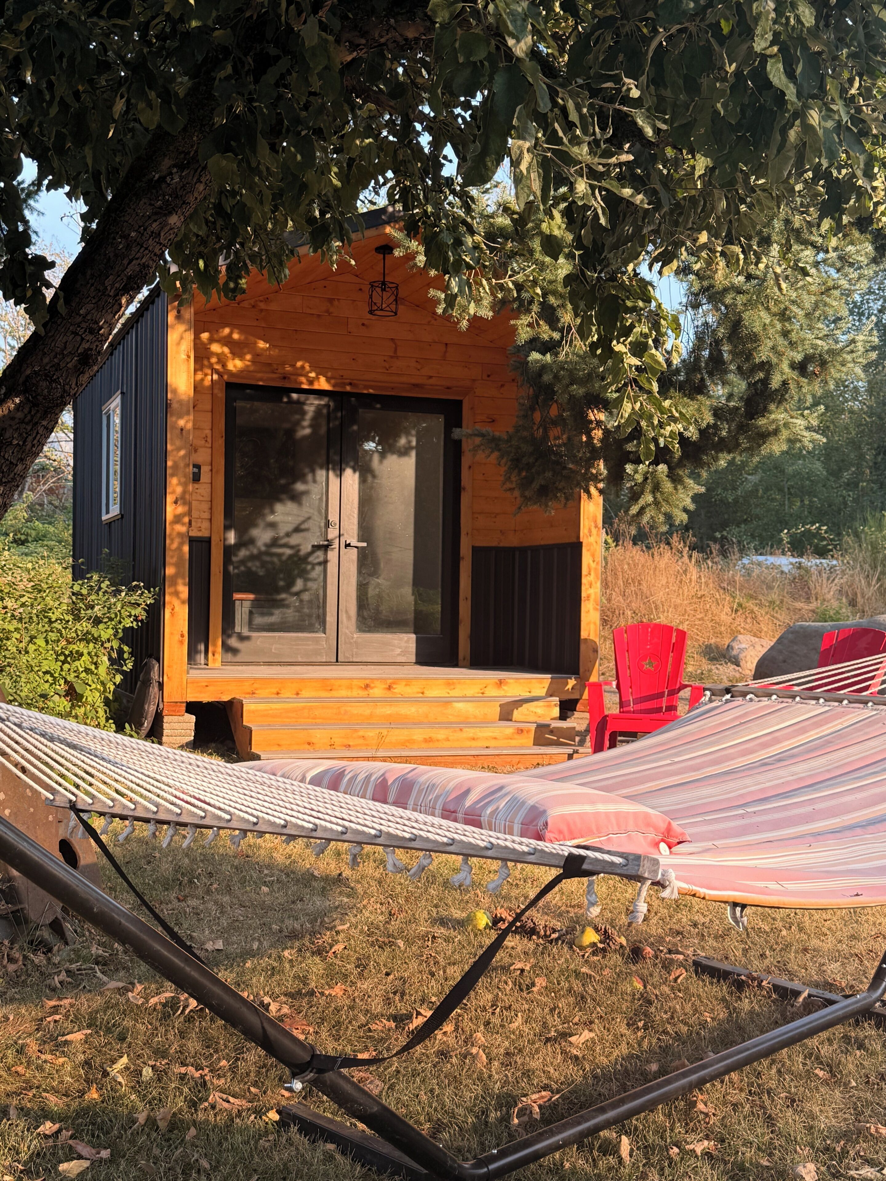 A wooden cabin with a large window and a door, a hammock, and a red chair.