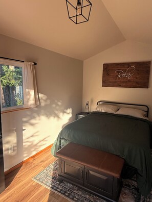 Un dormitorio con cama, una banca, una mesita de noche, una lámpara de pared y un ventanal con cortinas.