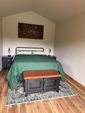 Un dormitorio con cama, mesitas de noche, un banco, una alfombra y una obra de arte en la pared.