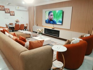 Een moderne woonkamer met een flatscreen televisie, beige banken, oranje fauteuils, een eethoek en een open haard.
