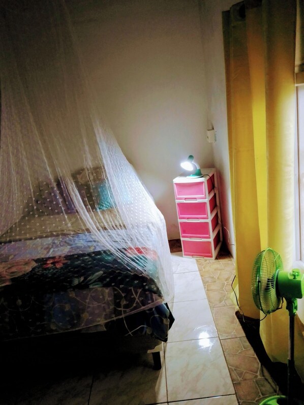 A bedroom with a bed covered by a mosquito net, a bedside table with a lamp, and a standing fan.