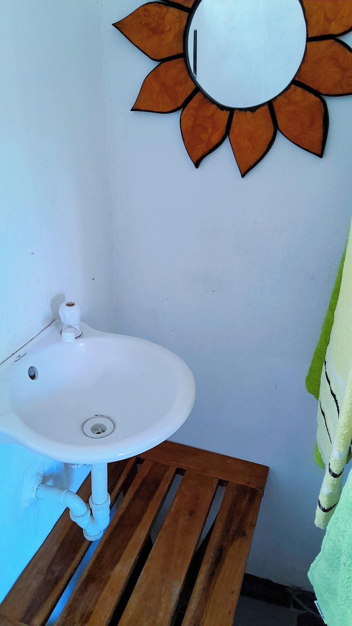 Une salle de bain avec un lavabo blanc, un banc en bois à lattes et un miroir mural orné d’un motif solaire.