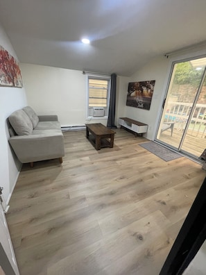 Een woonkamer met een grijze bank, een houten salontafel en een zitbankje. Er is een raam met zonwering en uitzicht op het omliggende landschap.