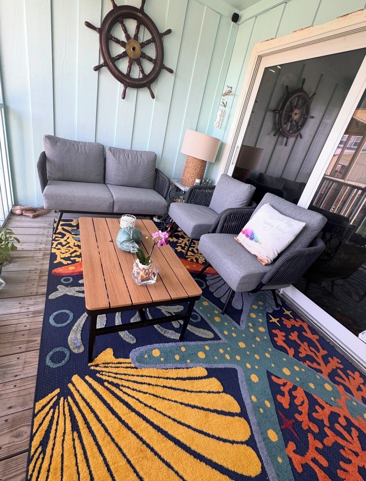 A cozy outdoor seating area with a sofa, chairs, and a table, featuring a decorative rug and wall-mounted ship wheel.