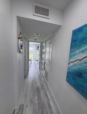 Un couloir avec un plancher en bois, des murs blancs, une bouche d’aération au plafond, un miroir rond et un tableau représentant une scène de mer.