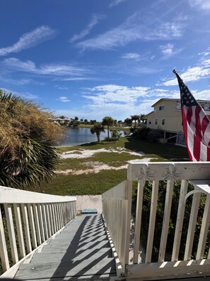 A wooden deck leading to a body of water with an American flag, a house, and palm trees in the background. - Don’t Worry, Beach Happy!  Little Gasparilla Island Vacation! Reserve now!! New! (Placida)